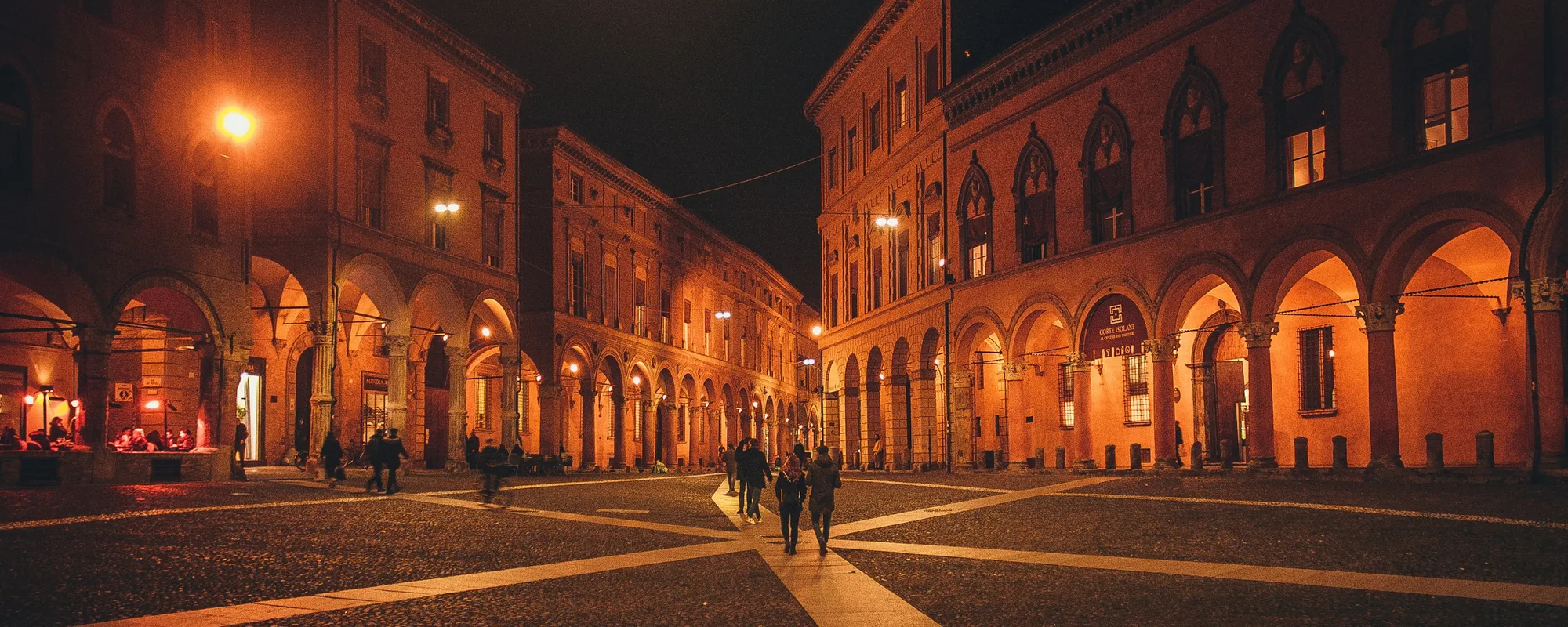 Arcaded palazzi glowing orange at night around a cobbled Bologna square, with people walking across it