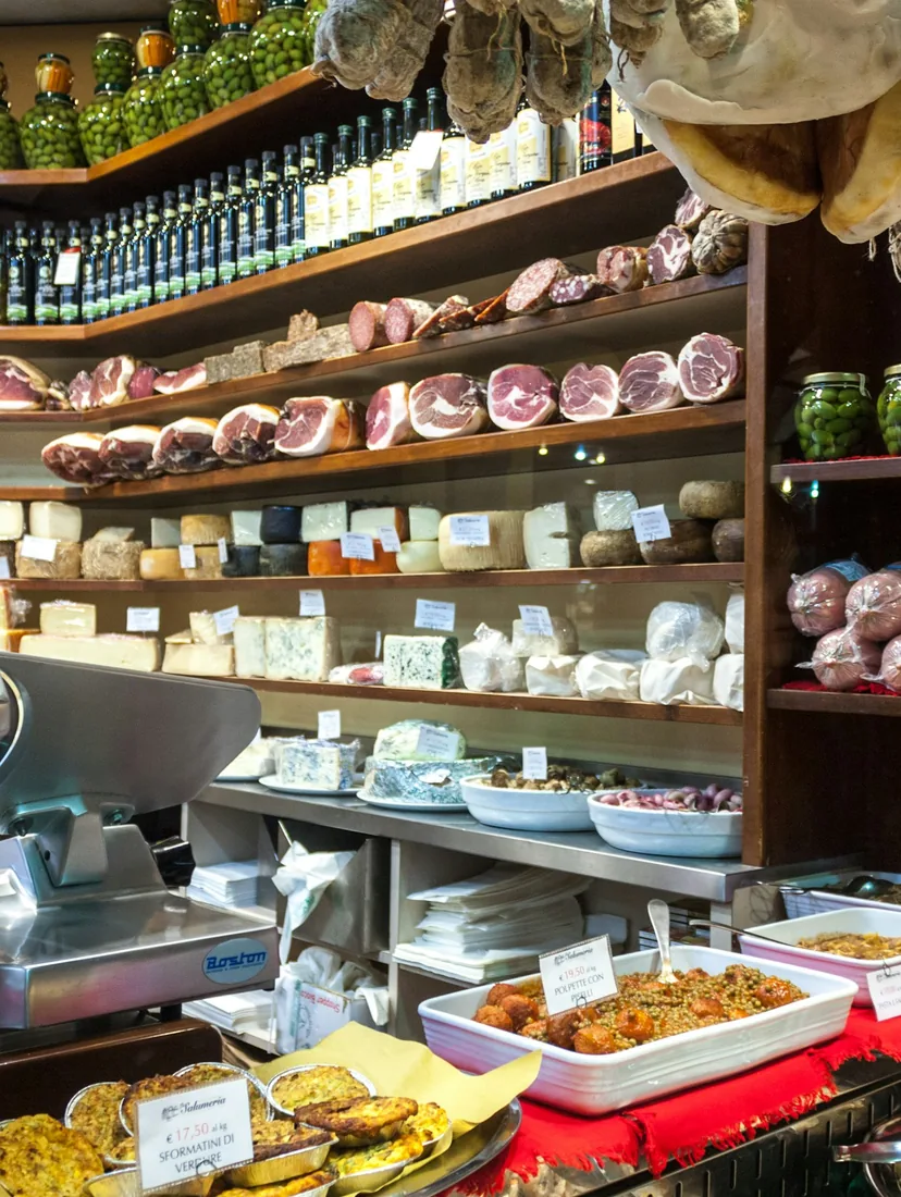 A delicatessen wall of cured hams, cheeses, jars and trays of prepared dishes behind a slicing machine