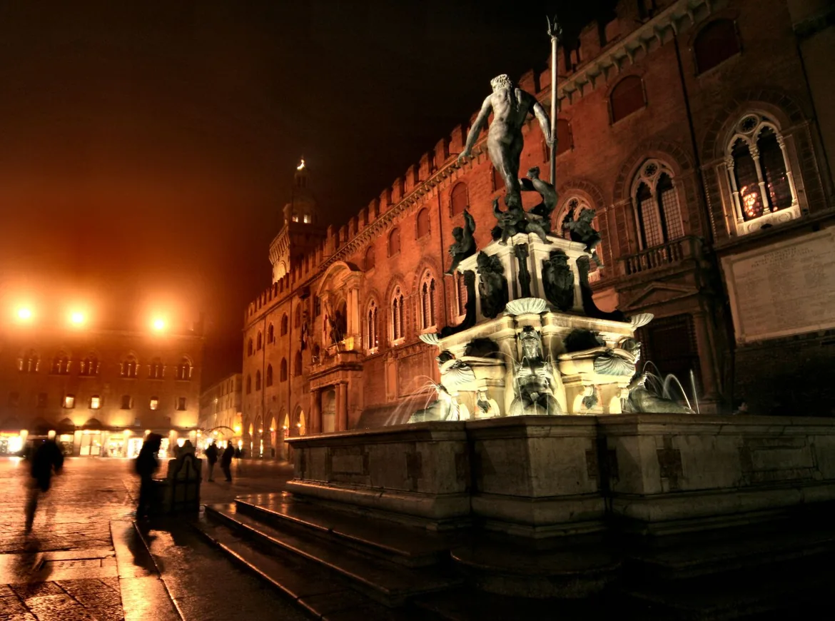 The Neptune Fountain lit up at night in front of a palace with arched windows, with figures crossing the square