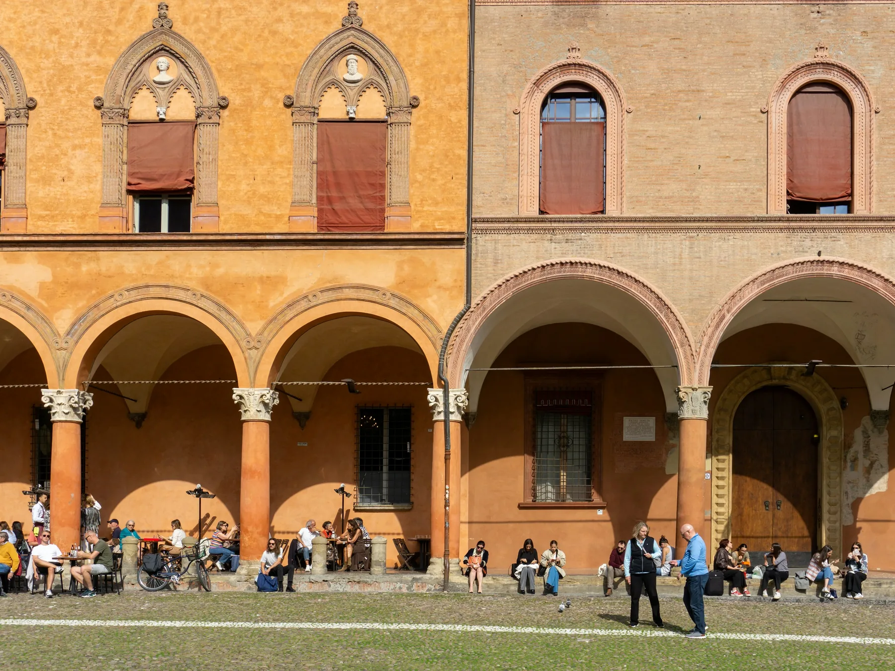 A row of round arches in ochre and pink stone with people sitting on the grass in front of them